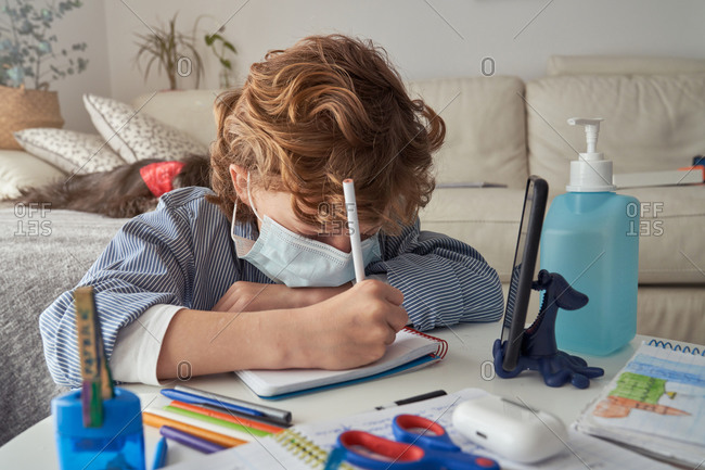 Boy in medical mask making notes in notepad while sitting at table with smartphone and disinfectant and studying online during quarantine