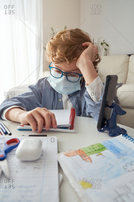Exhausted smart boy in glasses and medical mask doing homework assignment while sitting at table and studying remotely during quarantine at home