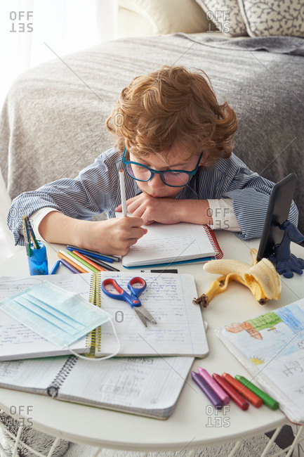 Boy in medical mask making notes in notepad while sitting at table with smartphone and disinfectant and studying online during quarantine