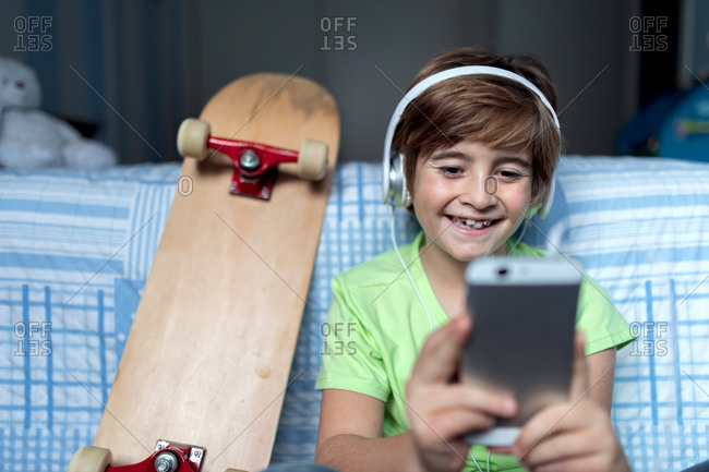Laughing little boy with headphones listening to music and chatting with friends in social network while sitting near skateboard in bedroom
