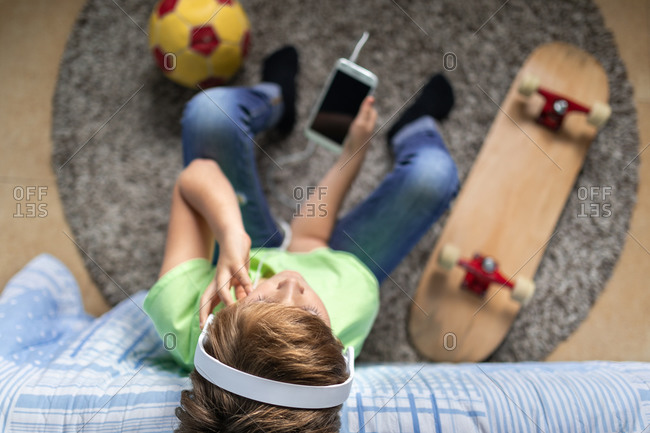 little boy with headphones listening to music and chatting with friends in social network while sitting near skateboard in bedroom