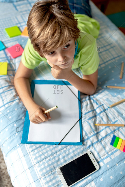 From above of thoughtful little boy in casual wear lying on bed with smartphone placed nearby and drawing on paper sheet with colorful pencils while spending time at home