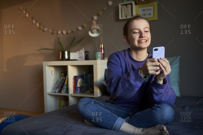Portrait of grinning girl sitting on bed taking selfie with smartphone