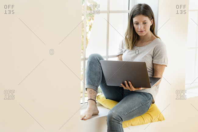 Young brunette woman at home sitting on window bench and holding her laptop working in home office