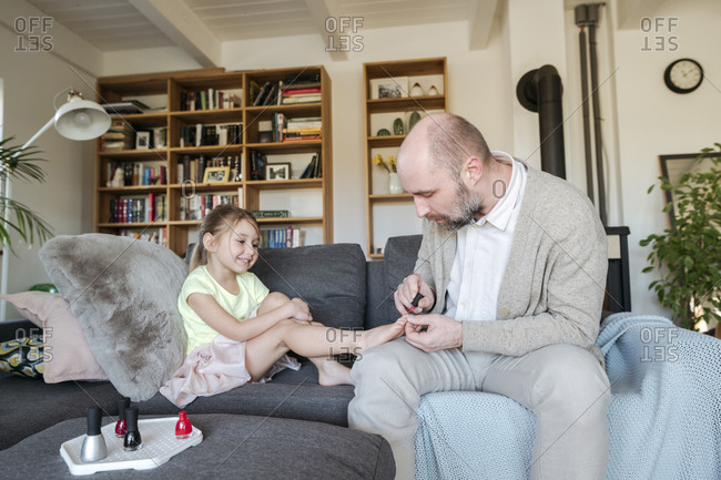 Father painting his daughter's toenails with red nail polish on the couch in living room