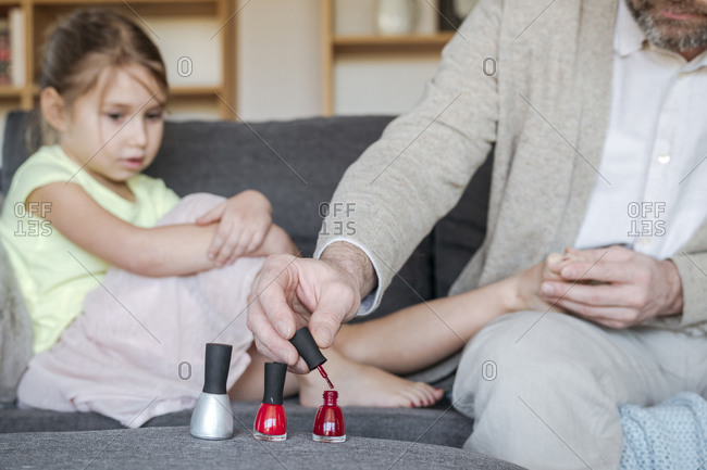 Crop view of father painting his daughter's toenails with red nail polish on the couch in living room