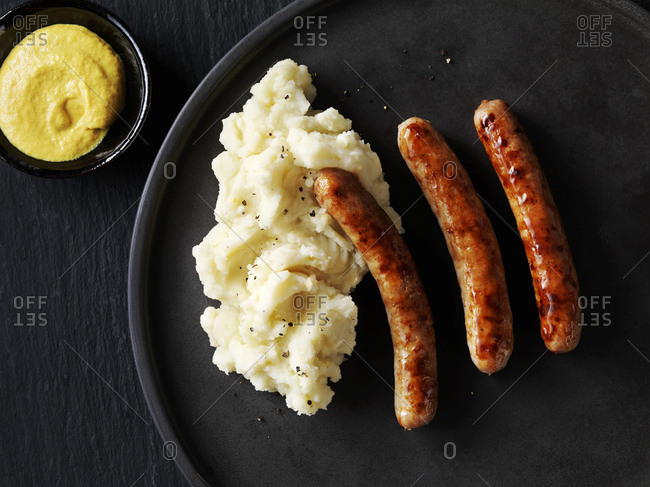 Still life with sausages and mash on black plate with bowl of  mustard, overhead view