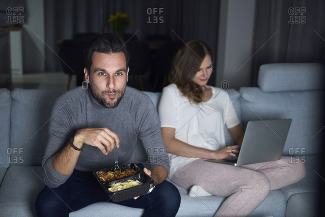 Young couple sitting on sofa in evening eating takeaway and using laptop