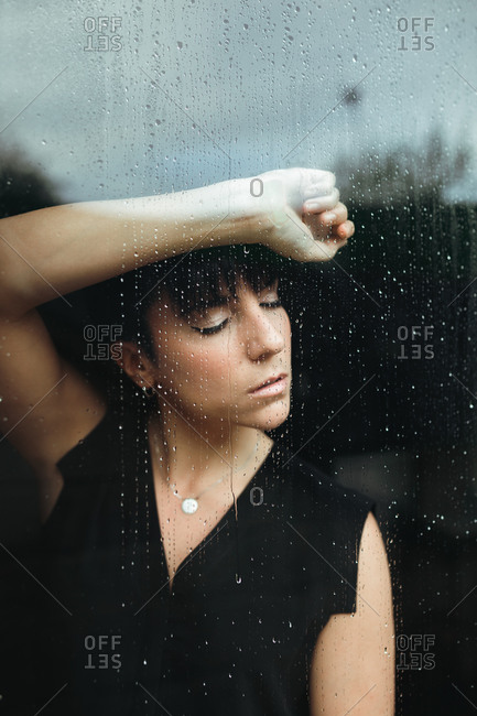 Portrait of a sad and nostalgic woman at home holding herself on a glass door with rain drops