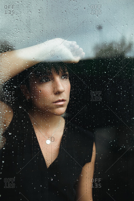 Portrait of a sad and nostalgic woman at home holding herself on a glass door with rain drops looking outside