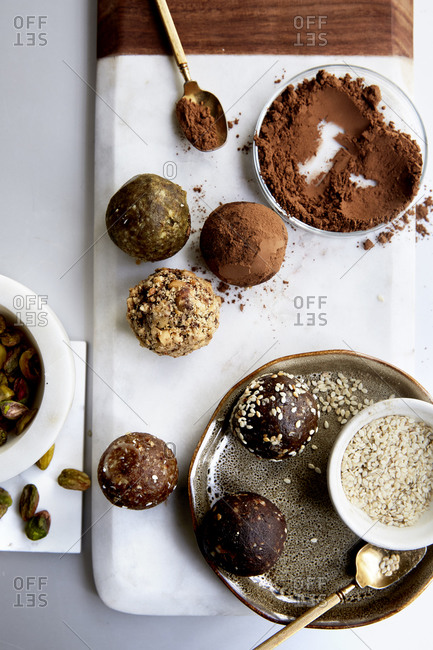 Date balls on a marble plate with cacao powder, sesame seeds and pistachios on the side on a white countertop,