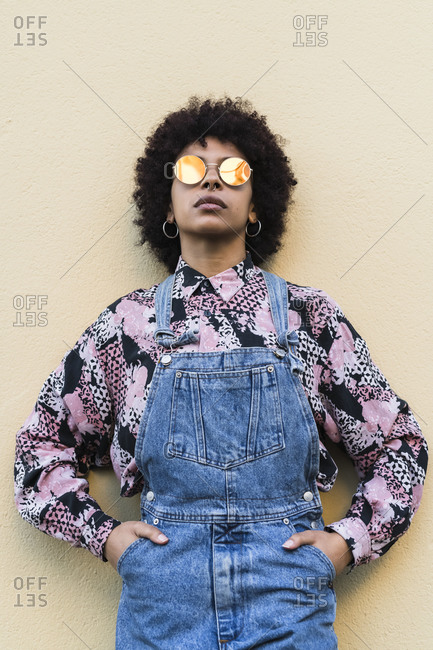 Cool young woman in dungarees and sunglasses standing in front of wall, portrait