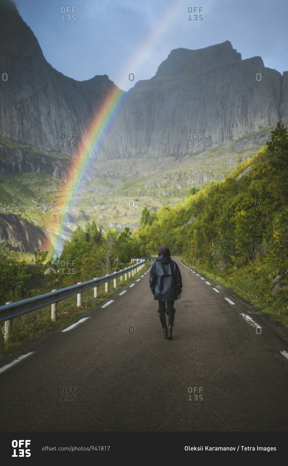 Norway, Lofoten Islands, Man walking down road with mountains and rainbow in background