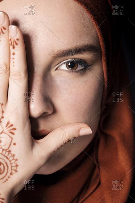 Portrait of a young adult Muslim female wearing brick orange hijab, half face covered with hand, close up