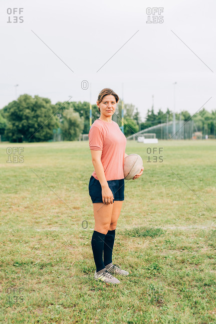 A woman standing on a training pitch in shorts and tee shirt holding a rugby ball.