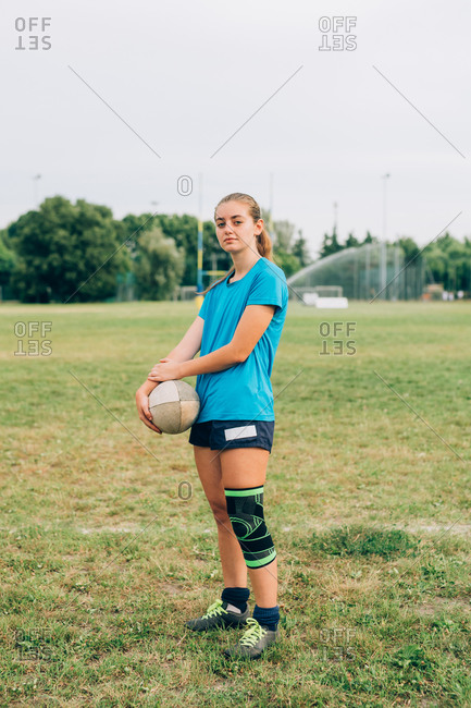 A woman standing on a training pitch in shorts and tee shirt with a knee strap holding a rugby ball.