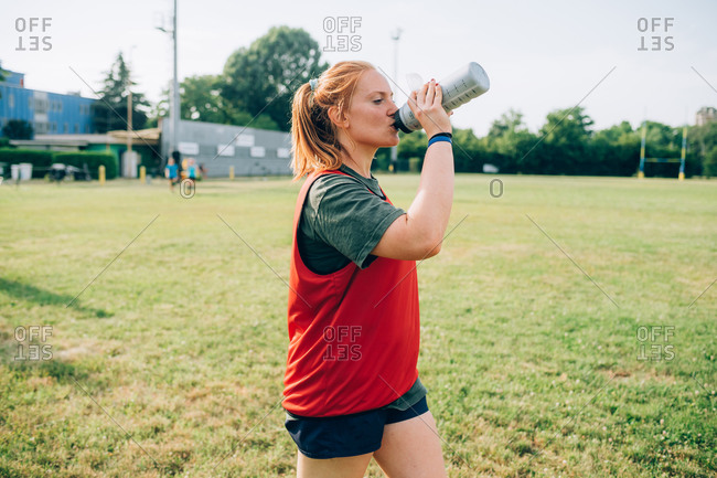 A woman walking across a training pitch drinking from a water bottle.