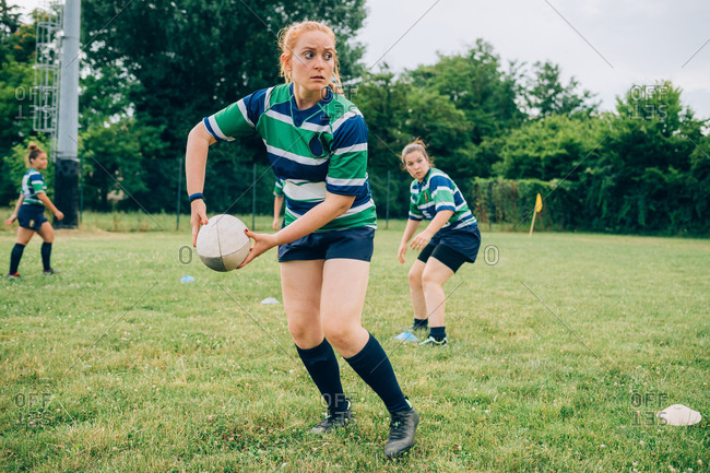 Three women wearing blue, white and green rugby shirts on a training pitch, one about to pass a rugby ball.