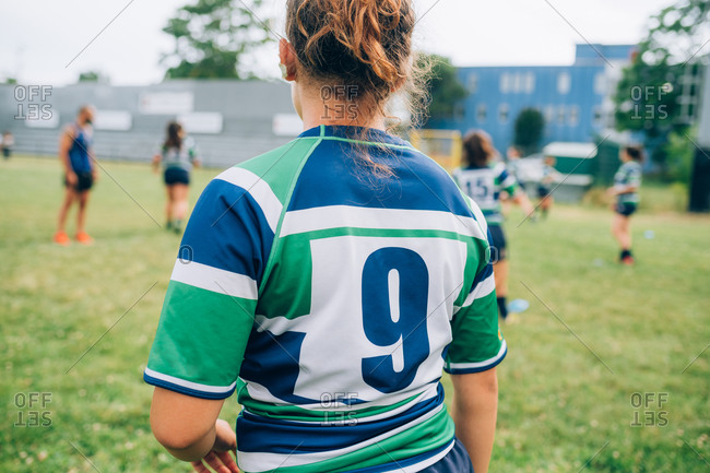 Rear view of a woman wearing a blue, green and white rugby shirt on a pitch at training with other players in the background.