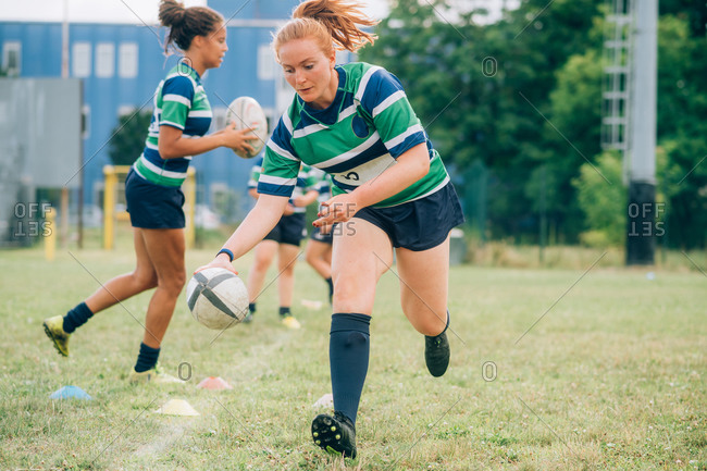 Three women wearing blue, white and green rugby shirts on a training pitch, one running with a rugby ball.