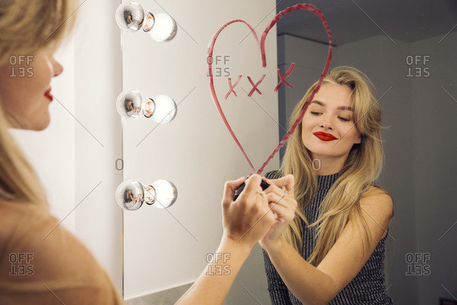 A woman looking in a bathroom mirror drawing a heart on the mirror with lipstick.