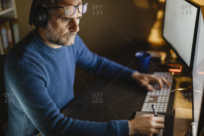 Mature man with headphones sitting at desk at home working with graphics tablet and computer