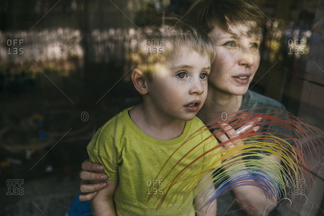 Portrait of mother and her little son looking out of window with drawn rainbow