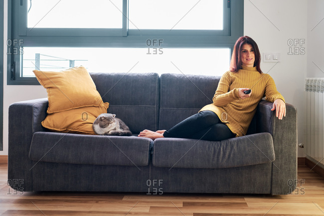 Woman on her couch using remote and watching tv with her exotic cat during corona virus outbreak