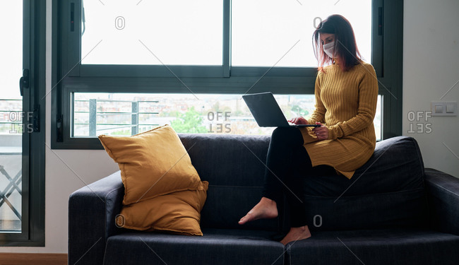 Woman working with laptop at home with mask on her couch on a cloudy day during coronavirus outbreak