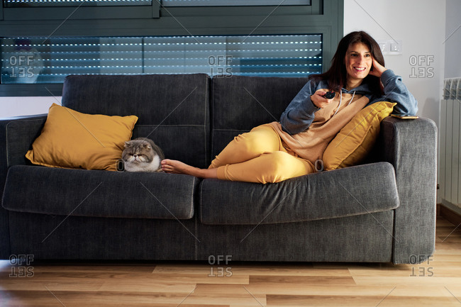 young woman on her couch with exotic cat using remote control while at home by corona virus