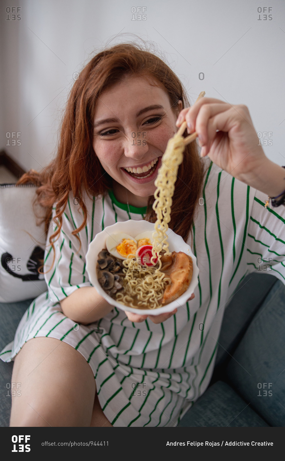 From above cheerful redhead female laughing and picking noodles from bowl of tasty ramen while sitting on couch at home