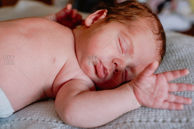Calm cute infant with closed eyes covered with blanket resting in bed against blurred interior of light bedroom