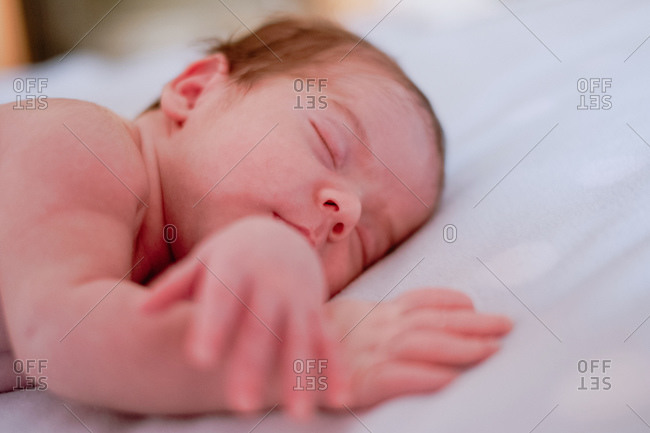 Calm cute infant with closed eyes covered with blanket resting in bed against blurred interior of light bedroom