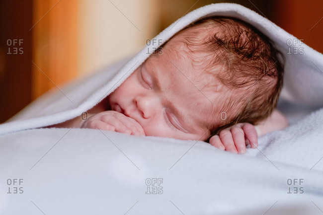 Calm cute infant with closed eyes covered with blanket resting in bed against blurred interior of light bedroom