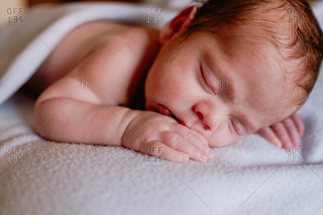 Calm cute infant with closed eyes covered with blanket resting in bed against blurred interior of light bedroom