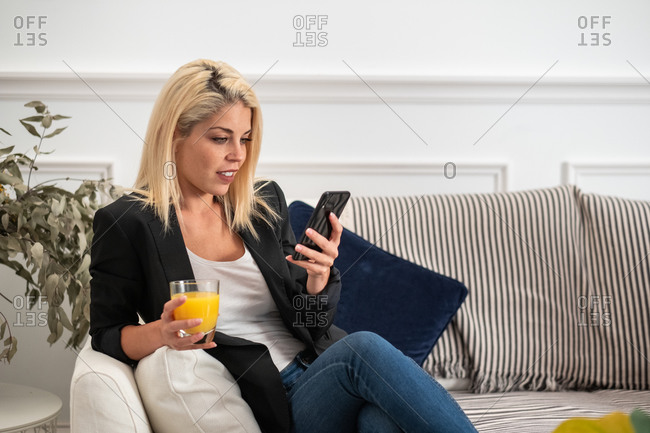 Happy blonde woman in casual clothes enjoying fresh orange juice and browsing smartphone while sitting on couch at home