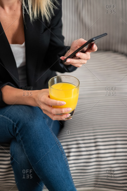Cropped unrecognizable blonde woman in casual clothes enjoying fresh orange juice and browsing smartphone while sitting on couch at home