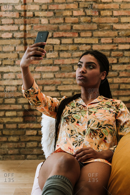 Serious young African American female in casual outfit taking selfie with smartphone while sitting on chair against brick wall in modern apartment