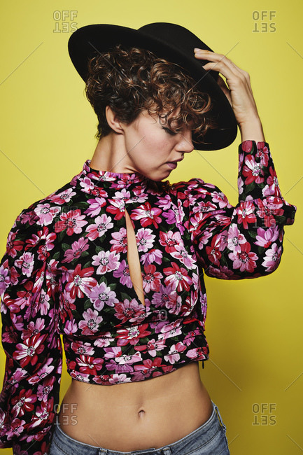 Fashionable happy female model holding in head a stylish black hat and colorful crop top with floral print standing against yellow background with closed eyes