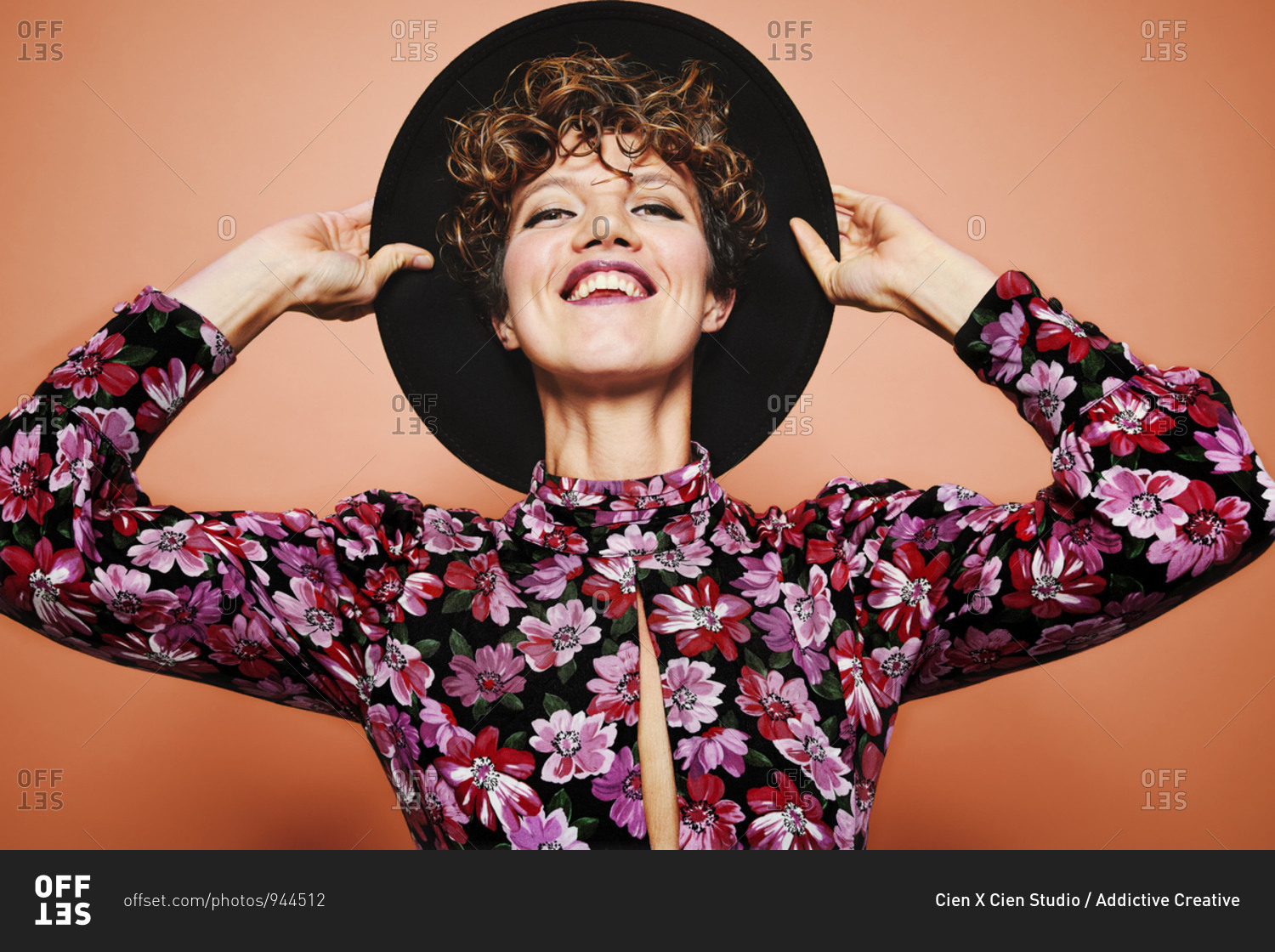 Optimistic young beautiful female model with curly hair wearing stylish black hat and colorful blouse looking at camera and smiling while standing against orange background