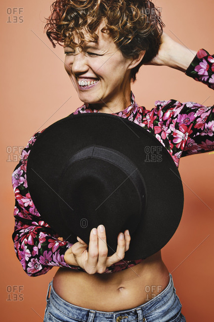 Fashionable happy female model holding a stylish black hat in colorful crop top with floral print standing against orange background with closed eyes