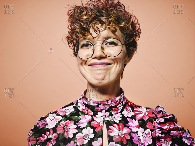 Happy young curly haired female in stylish floral blouse and trendy eyeglasses smiling and looking at camera against red background