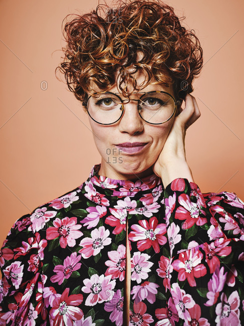 Thoughtful doubtful beautiful curly haired female in trendy eyeglasses and stylish colorful blouse with floral ornament looking at camera against pink background