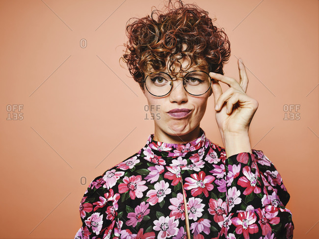 Thoughtful doubtful beautiful curly haired female in trendy eyeglasses and stylish colorful blouse with floral ornament looking at camera against pink background