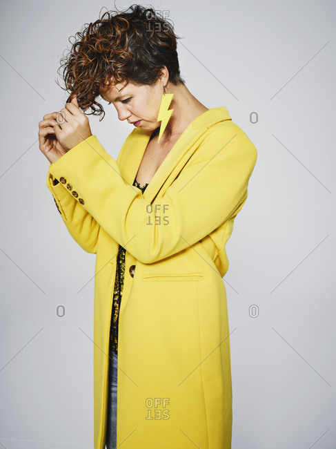 Adult female in stylish yellow coat and with lightning earring using comb to style curly hair against gray background