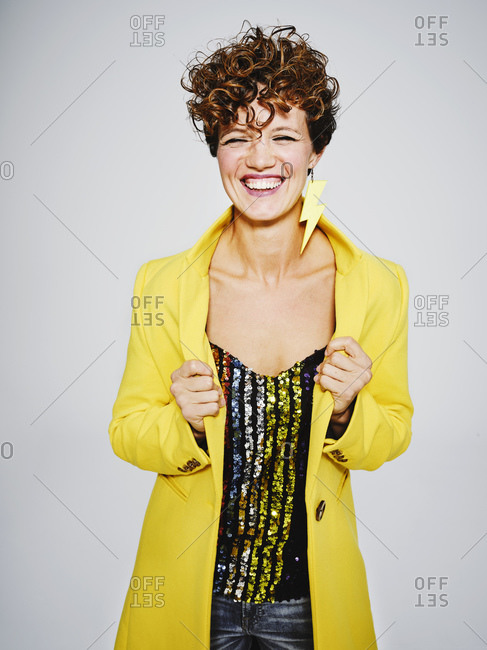 Cheerful woman with sequin top and lightning earring smiling and adjusting stylish yellow coat against gray background