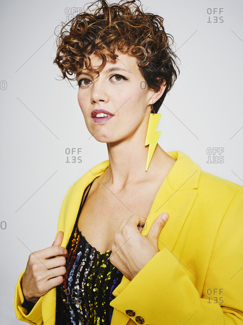 Portrait of cheerful woman with sequin top and lightning earring smiling and adjusting stylish yellow coat against gray background