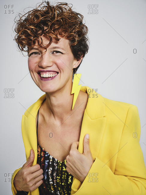 Portrait of cheerful woman with sequin top and lightning earring smiling and adjusting stylish yellow coat against gray background
