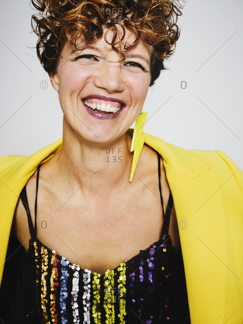 Portrait of cheerful woman with sequin top and lightning earring and stylish yellow coat smiling to the camera while standing against gray background