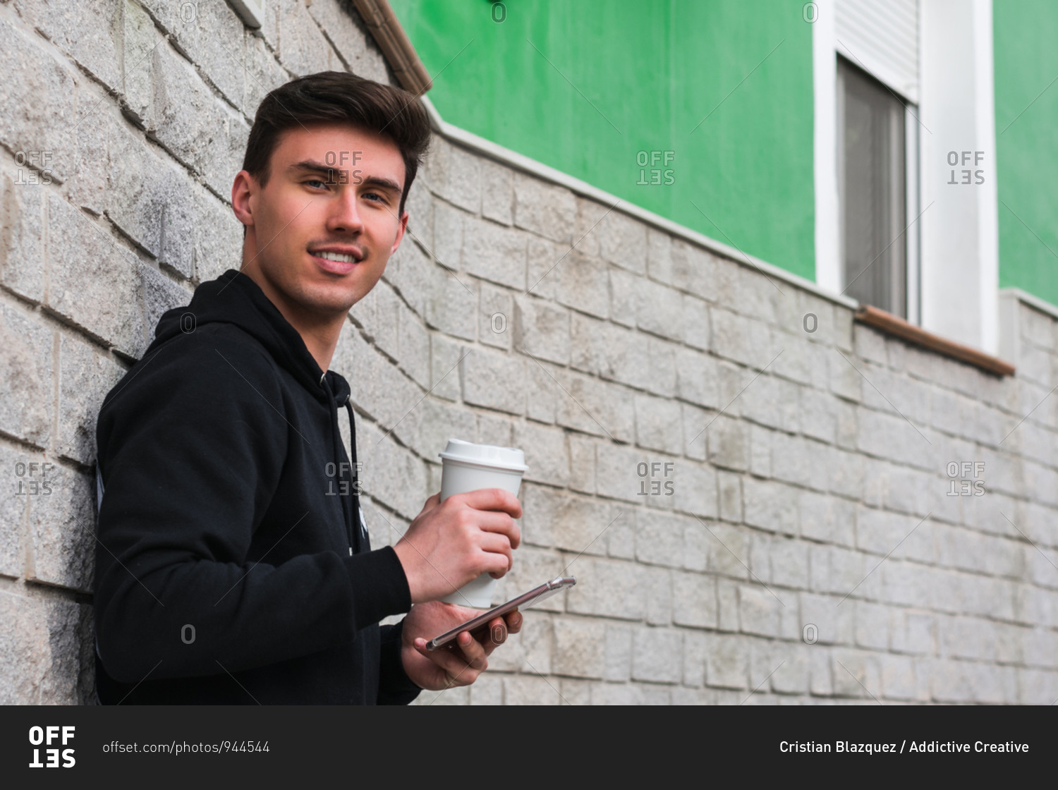 Side view of positive young male enjoying takeaway beverage and using smartphone while leaning on building wall and looking at camera on city street